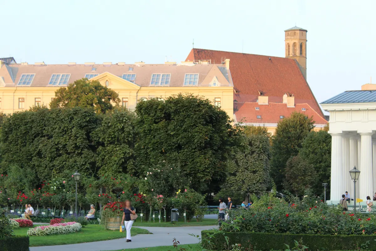 The Minoritenkirche seen across green gardens from the Volksgarten in Vienna, with its tower and red roof rising behind a low yellow building